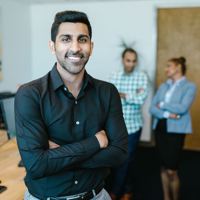 A finance professional standing in an office with colleagues talking behind him